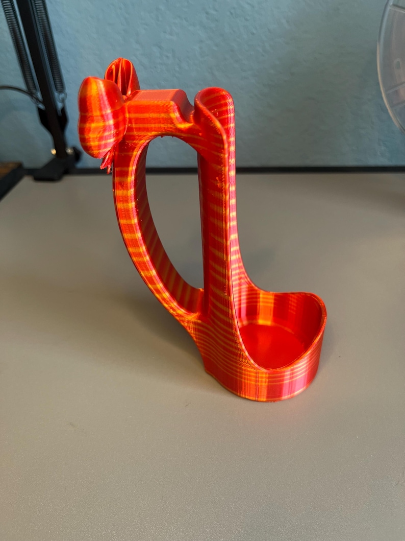 Puede incluir: Un soporte para tel&eacute;fono impreso en 3D, a rayas rojas y amarillas. El soporte tiene un asa curva y una base peque&ntilde;a y redondeada para sostener un tel&eacute;fono. La parte superior tiene un dise&ntilde;o en forma de lazo. El soporte es de pl&aacute;stico.