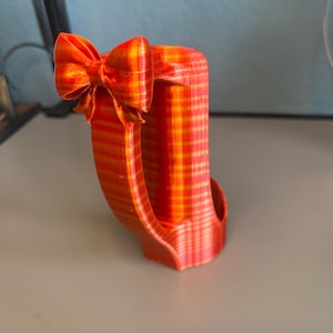 Puede incluir: Objeto decorativo impreso en 3D, a rayas naranjas y amarillas, con un lazo. El objeto tiene un asa curva y un cuerpo rectangular. El lazo est&aacute; en la parte superior izquierda.