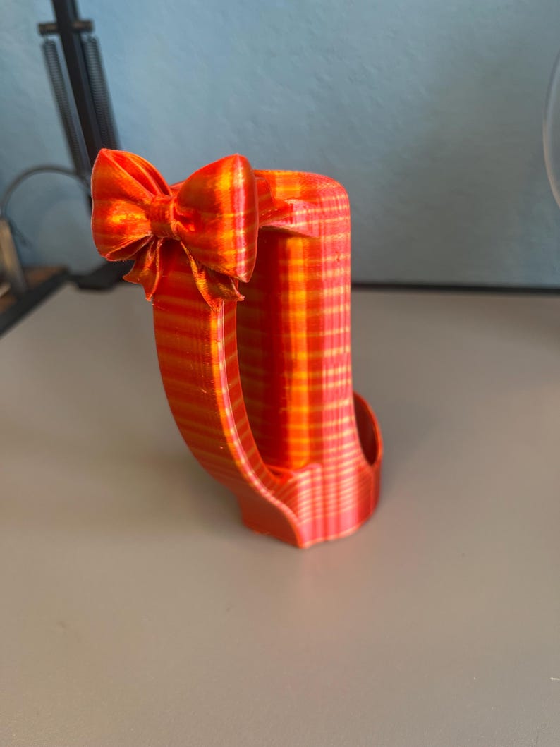 Puede incluir: Objeto decorativo impreso en 3D, a rayas naranjas y amarillas, con un lazo. El objeto tiene un asa curva y un cuerpo rectangular. El lazo est&aacute; en la parte superior izquierda.