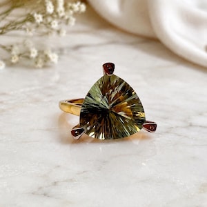 May include: A gold ring featuring a large, triangular-cut, faceted green gemstone. The stone displays a radial pattern of light and dark olive-green tones. The setting includes three small prongs that secure the gemstone to the polished gold band.
