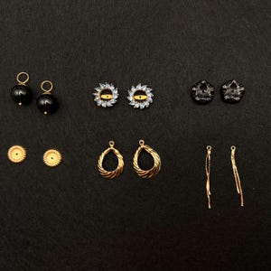May include: A collection of various earrings displayed on a black surface. The earrings include black pearl studs, gold-toned teardrop hoops, and delicate chain earrings. Other styles feature blue and clear crystal accents.