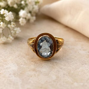 May include: A gold ring with an oval, light blue gemstone. The ring has a detailed gold band with a decorative design on the sides. The gemstone is set in a gold bezel.