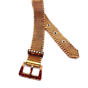 May include: A gold-toned mesh belt with a rectangular buckle adorned with red gemstones. The belt features a series of small, round embellishments along the edges and a row of pearls near the buckle. The belt is displayed against a white background.