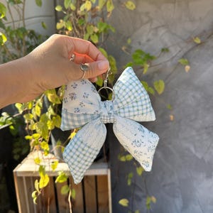 May include: A large, decorative bow keyring with a silver clasp. The bow is made of fabric with a mix of patterns, including small blue floral prints and a blue and white gingham check. The bow is held up by a hand.