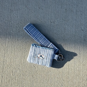 May include: A blue and white striped wristlet with a matching key fob. The wristlet features an embroidered design of a white and black robot head with stars. The key fob has a silver key ring.