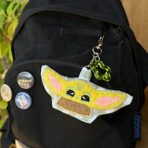 May include: Black backpack with a zip, adorned with a Baby Yoda keyring and pin badges. The keyring includes a green star-shaped charm. The Baby Yoda charm is yellow, brown, and white.
