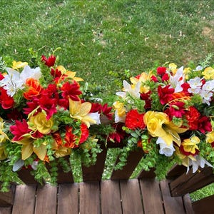May include: Two floral arrangements featuring red, yellow and white flowers. The arrangements are positioned on a wooden railing with green grass in the background.