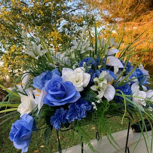 May include: A blue and white artificial flower arrangement featuring lilies, roses, and carnations. The arrangement is displayed on a black metal stand.