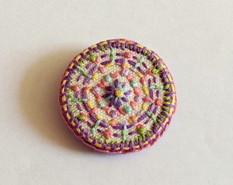 Hand embroidered circular fabric brooch. Pink and purple pattern with pops of blue, green and yellow.