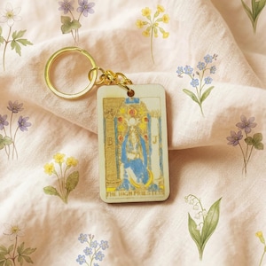 Op de afbeelding: Een rechthoekige sleutelhanger met een tarotkaartontwerp en goudkleurige hardware. De kaart toont een figuur in blauw en goud, met de woorden "THE HIGH PRIESTESS". De achtergrond is een lichtroze stof met bloemenillustraties.