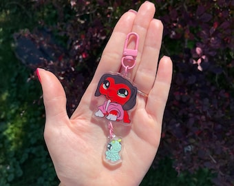 以下が含まれることがあります： ピンク色のキーホルダー。ロブスタークラスプで2つのアクリルチャームを保持しています。上部のチャームはピンク色の服を着た赤い犬です。下部のチャームは水色のカエルです。背景は緑と紫の葉のぼやけた景色です。