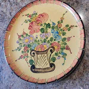 May include: A round ceramic plate with a hand-painted floral design. The plate features a yellow background with a pink, blue, and green floral bouquet in a brown vase. The rim of the plate is decorated with a pink and black stripe.