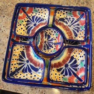 Könnte beinhalten: Dekorative Serviertablett mit einem quadratischen Design und einem tiefblauen Rand. Das Tablett hat eine zentrale runde Schale und vier quadratische Schalen, alle mit einem farbenfrohen Blumendesign in Blau, Rot, Gelb und Orange. Aus Keramik.