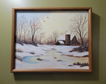 May include: Framed landscape painting depicting a winter scene with a snow-covered barn and silo. Bare trees line the horizon, and a frozen stream flows through the foreground. The artwork is encased in a wooden frame.