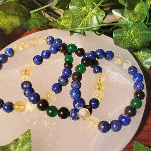 May include: Three beaded bracelets with blue, green, and yellow beads. The bracelets are arranged on a white surface.