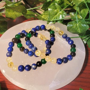 May include: Three beaded bracelets with blue, green, and yellow beads. The bracelets are stacked on top of each other on a white surface.