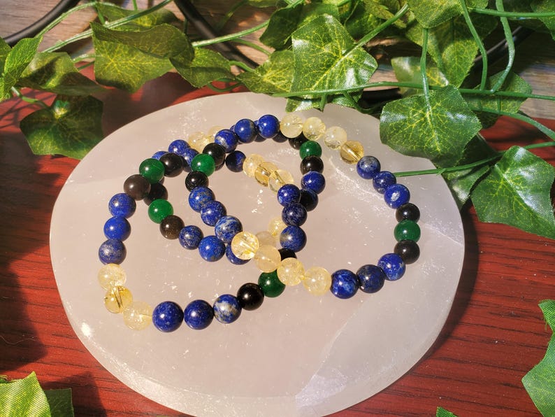 May include: Three beaded bracelets with blue, green, and yellow beads. The bracelets are stacked on top of each other on a white surface.
