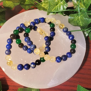 May include: Three beaded bracelets with blue, yellow, green, and black beads. The bracelets are on a white surface.