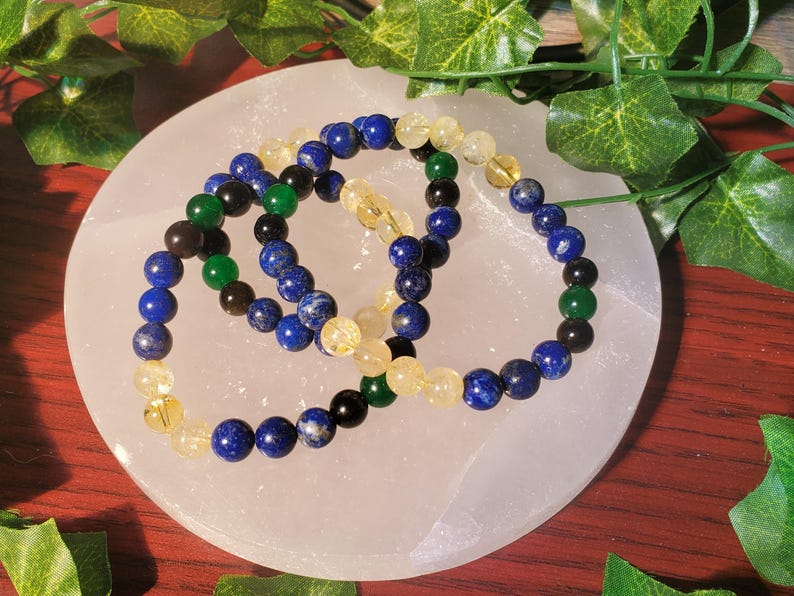 May include: Three beaded bracelets with blue, yellow, green, and black beads. The bracelets are on a white surface.
