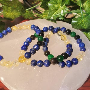 May include: Three beaded bracelets with blue, yellow, green, and black beads. The bracelets are on a white surface.