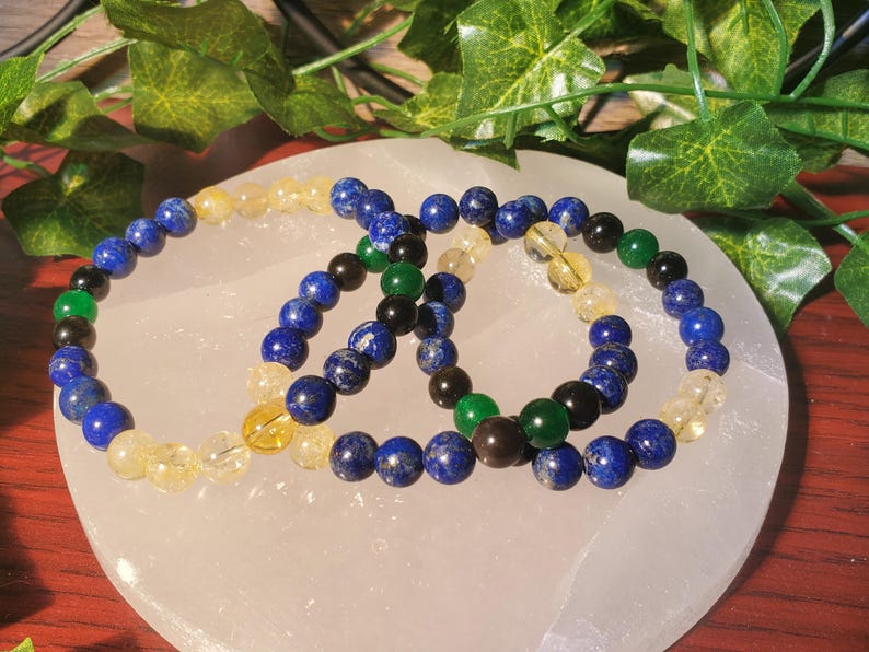 May include: Three beaded bracelets with blue, yellow, green, and black beads. The bracelets are on a white surface.