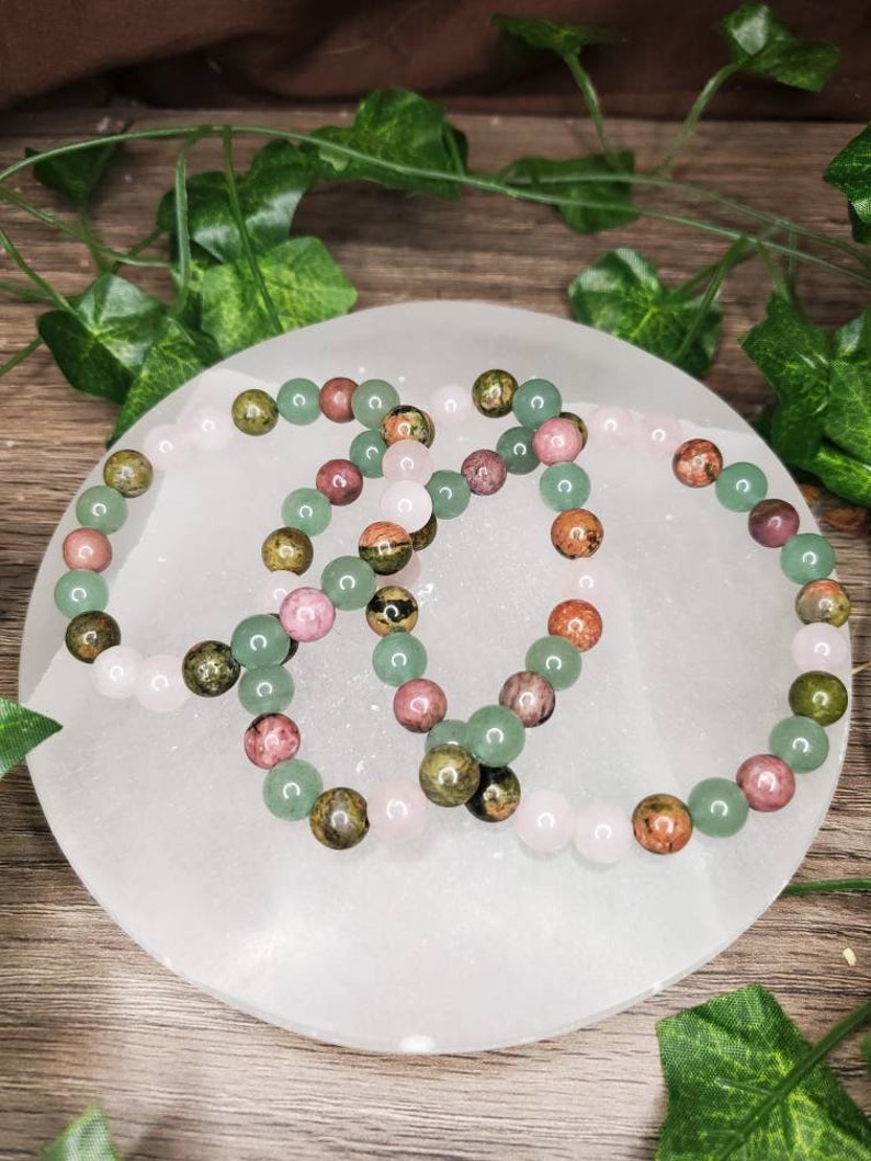 Peut inclure: Trois bracelets en perles compos&eacute;es d'aventurine verte, de jaspe rose et vert et de quartz rose. Les bracelets sont dispos&eacute;s en cercle sur une plaque de recharge en s&eacute;l&eacute;nite blanche.