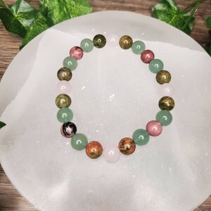Peut inclure: Un bracelet compos&eacute; de perles de pierres pr&eacute;cieuses vertes, roses et brunes. Le bracelet est sur une surface blanche.