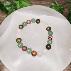 Peut inclure: Un bracelet compos&eacute; de perles de pierres pr&eacute;cieuses vertes, roses et brunes. Le bracelet est sur une surface blanche.