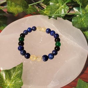 May include: A bracelet made of blue, green, black, and yellow beads. The bracelet is on a white surface.