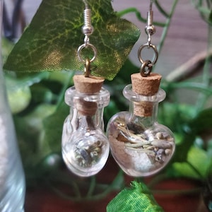 May include: A pair of silver heart-shaped glass bottle earrings with cork stoppers and silver hooks. The bottles are filled with dried herbs and flowers.