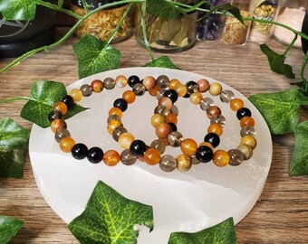 May include: Three stacked bracelets made of carnelian, smoky quartz, and black onyx beads. The bracelets are on a white selenite charging plate.