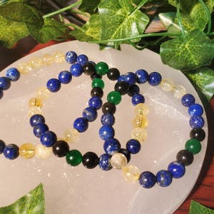 May include: Three beaded bracelets with blue, green, and yellow beads. The bracelets are arranged on a white surface.