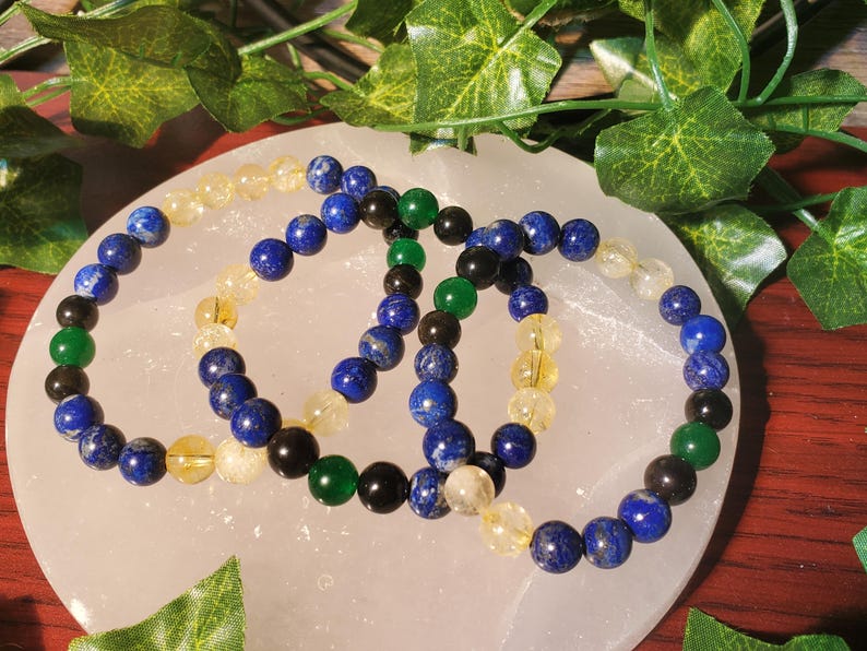 May include: Three beaded bracelets with blue, green, and yellow beads. The bracelets are arranged on a white surface.