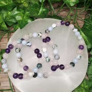 May include: Two bracelets made with amethyst, rose quartz, and moss agate beads. The bracelets are on a white circular surface.