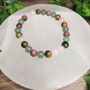 Peut inclure: Un bracelet compos&eacute; de perles de pierres pr&eacute;cieuses vertes, roses et brunes. Le bracelet est sur une plaque de recharge en s&eacute;l&eacute;nite blanche.