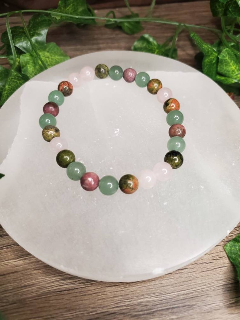 Peut inclure: Un bracelet compos&eacute; de perles de pierres pr&eacute;cieuses vertes, roses et brunes. Le bracelet est sur une plaque de recharge en s&eacute;l&eacute;nite blanche.