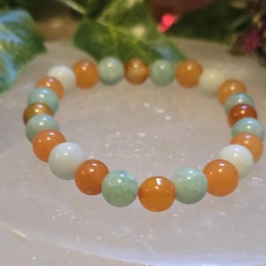 May include: A beaded bracelet featuring a mix of orange, white, and light green round beads. The bracelet is made of polished stones and is displayed on a white surface. The bracelet is a piece of jewelry.