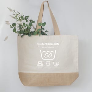 May include: A beige tote bag with a natural jute bottom and handles. The bag features white text and laundry symbols, including a 50-degree wash symbol. Greenery is visible inside the bag.