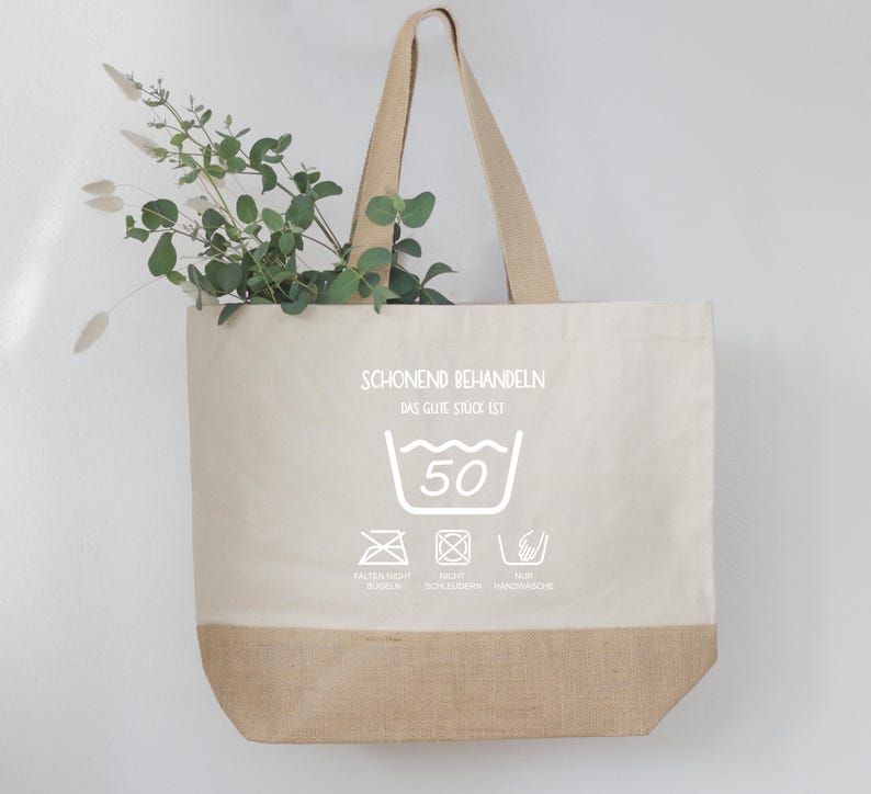 May include: A beige tote bag with a natural jute bottom and handles. The bag features white text and laundry symbols, including a 50-degree wash symbol. Greenery is visible inside the bag.