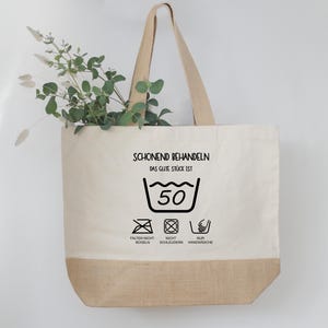 May include: A beige tote bag with a brown bottom and handles. The bag features the text "SCHONEND BEHANDELN" and washing instructions, including a 50-degree wash symbol. Greenery is placed inside the bag.