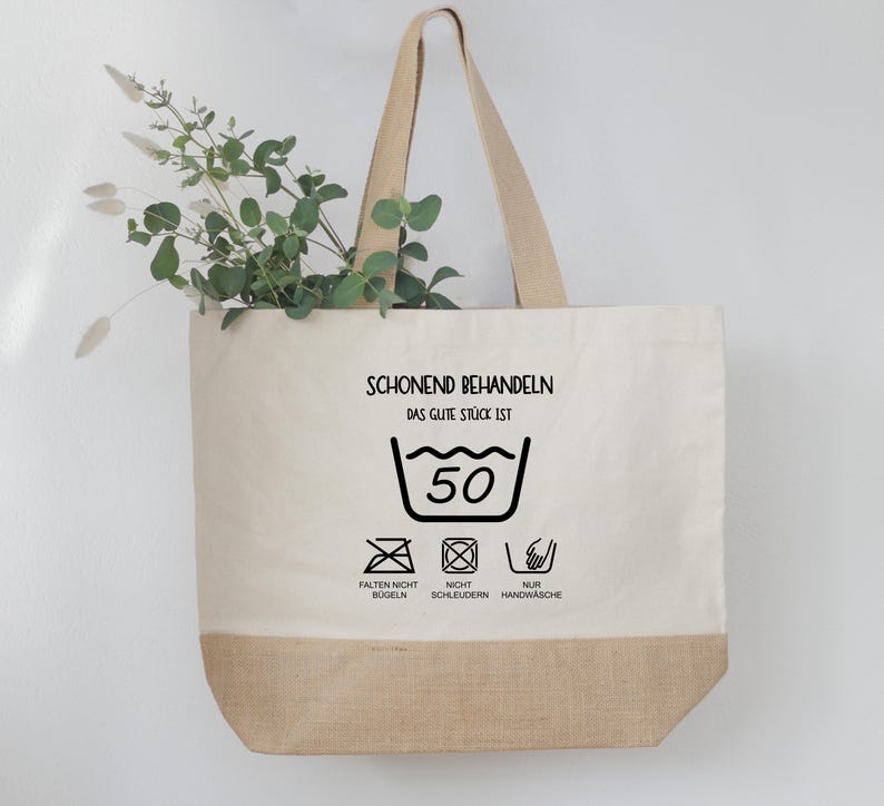May include: A beige tote bag with a brown bottom and handles. The bag features the text "SCHONEND BEHANDELN" and washing instructions, including a 50-degree wash symbol. Greenery is placed inside the bag.