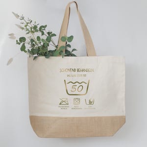 May include: A beige tote bag with a natural burlap bottom and handles. The bag features gold text and washing symbols, with a sprig of greenery inside. The text reads "SCHONEND BEHANDELN" and "DAS GUTE ST&Uuml;CK IST".