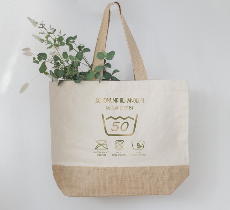 May include: A beige tote bag with a natural burlap bottom and handles. The bag features gold text and washing symbols, with a sprig of greenery inside. The text reads "SCHONEND BEHANDELN" and "DAS GUTE ST&Uuml;CK IST".