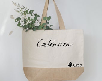 May include: A white canvas tote bag with brown burlap trim. The bag has the text "Catmom" in black cursive script and a black paw print with the text "Oreo" below it.