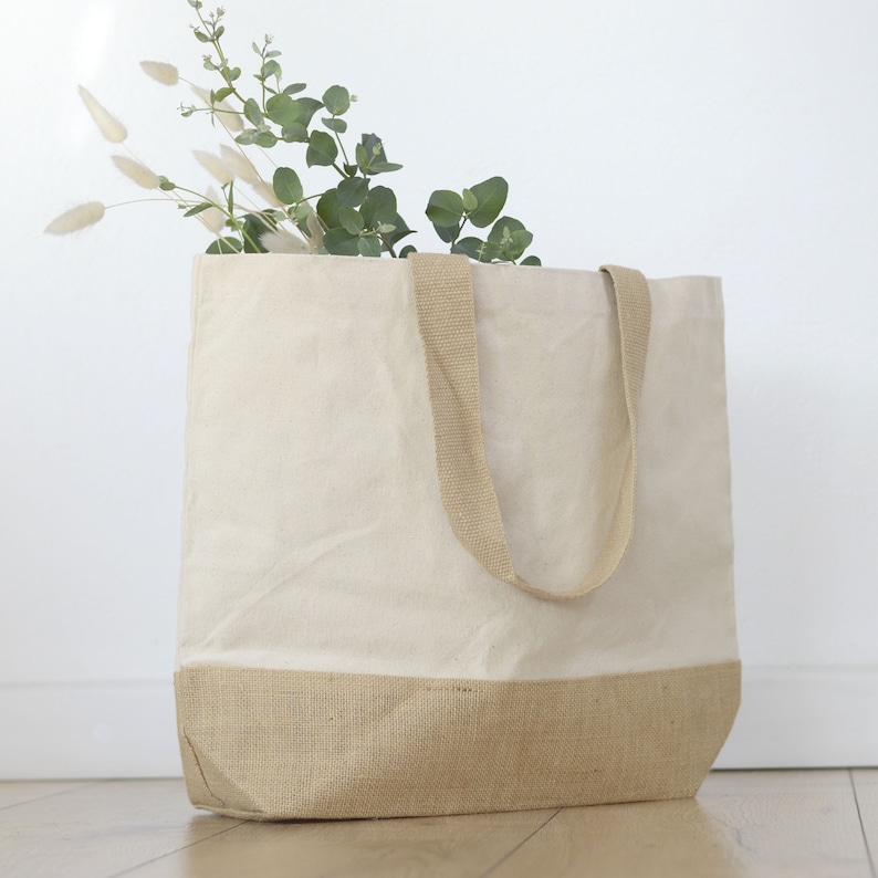 May include: A large, beige canvas tote bag with a natural burlap bottom and matching handles. The bag is filled with green eucalyptus and white decorative grasses, creating a natural, minimalist aesthetic.