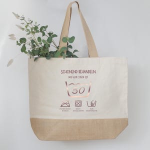 May include: A beige tote bag with a brown bottom and handles. The bag features text and washing symbols in a rose gold color. Greenery is visible inside the bag.