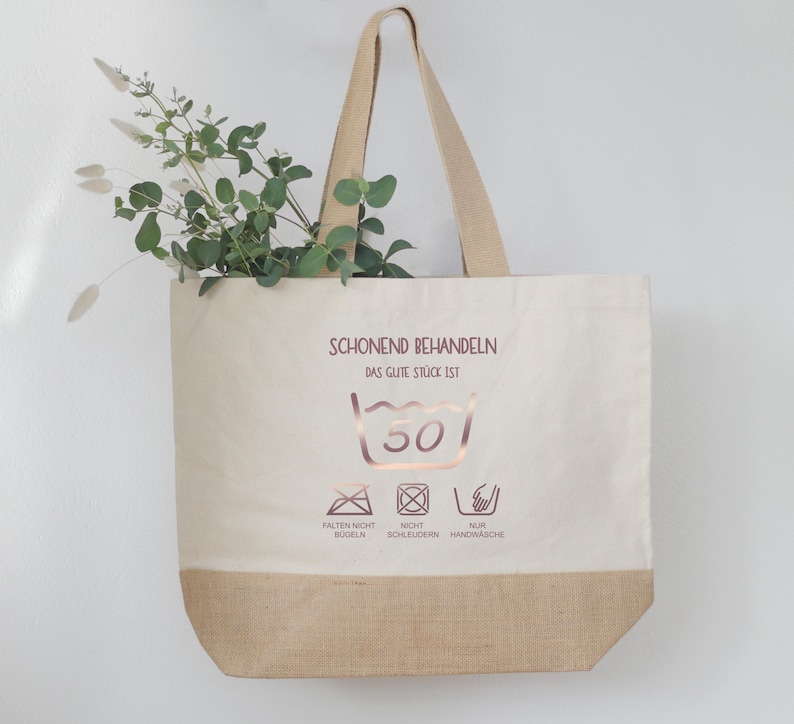 May include: A beige tote bag with a brown bottom and handles. The bag features text and washing symbols in a rose gold color. Greenery is visible inside the bag.