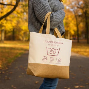 May include: A beige tote bag with a brown bottom section and matching handles. The bag features text and laundry symbols, including a 50-degree Celsius wash symbol. The bag is being held in front of a blurred autumn background.