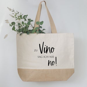 Könnte beinhalten: Eine beigefarbene Tragetasche mit einem Boden und Griffen aus Jute. Auf der Tasche steht in schwarzer Schrift "zu Vino sag ich nie no!". Grünzeug ist im Inneren der Tasche zu sehen.
