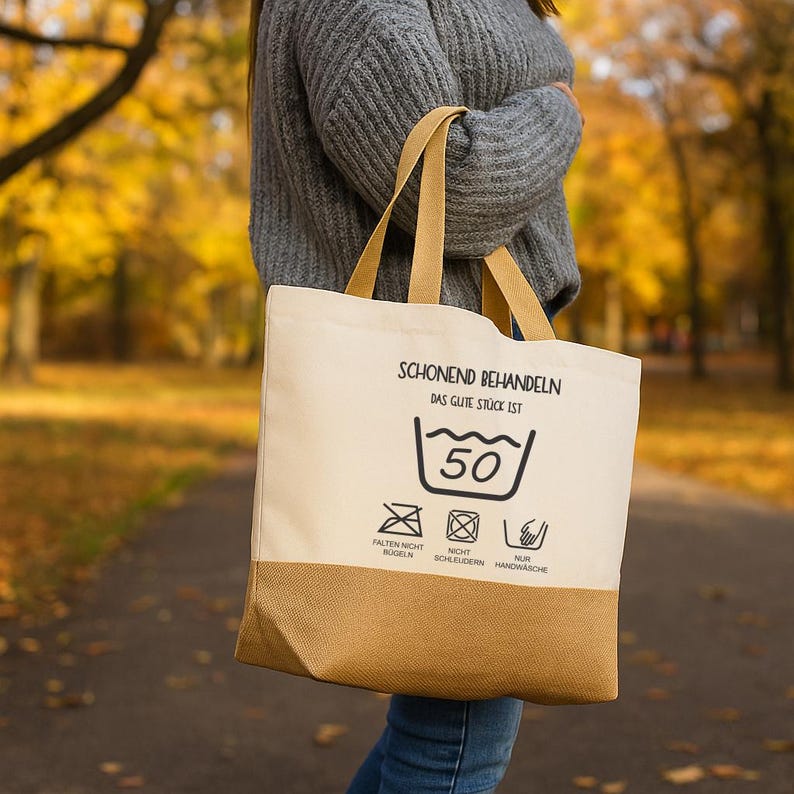 May include: A beige and tan tote bag with a printed laundry care label. The label includes text and symbols indicating washing instructions. The bag has tan handles and a tan bottom section.